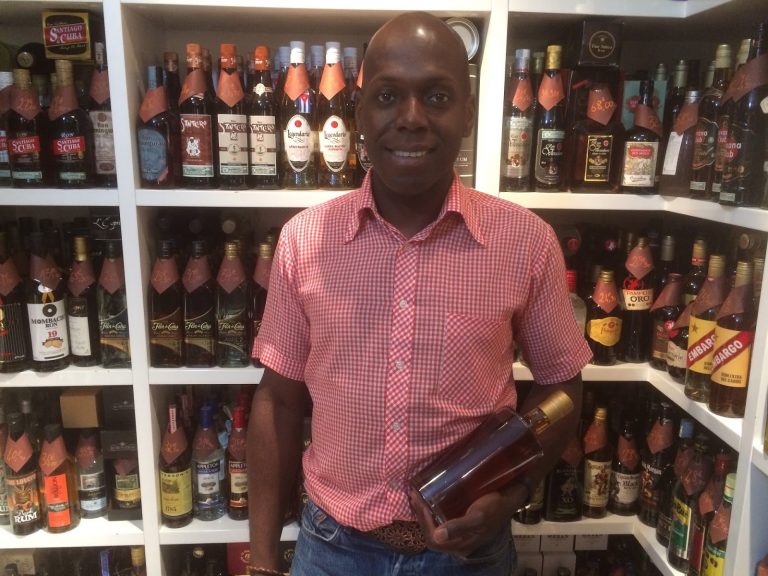 Photo de Freddy au centre, en chemise rose tenant une bouteille de rhum à la main avec en arrière plan les étales de bouteille de la boutique de À'Rhûm