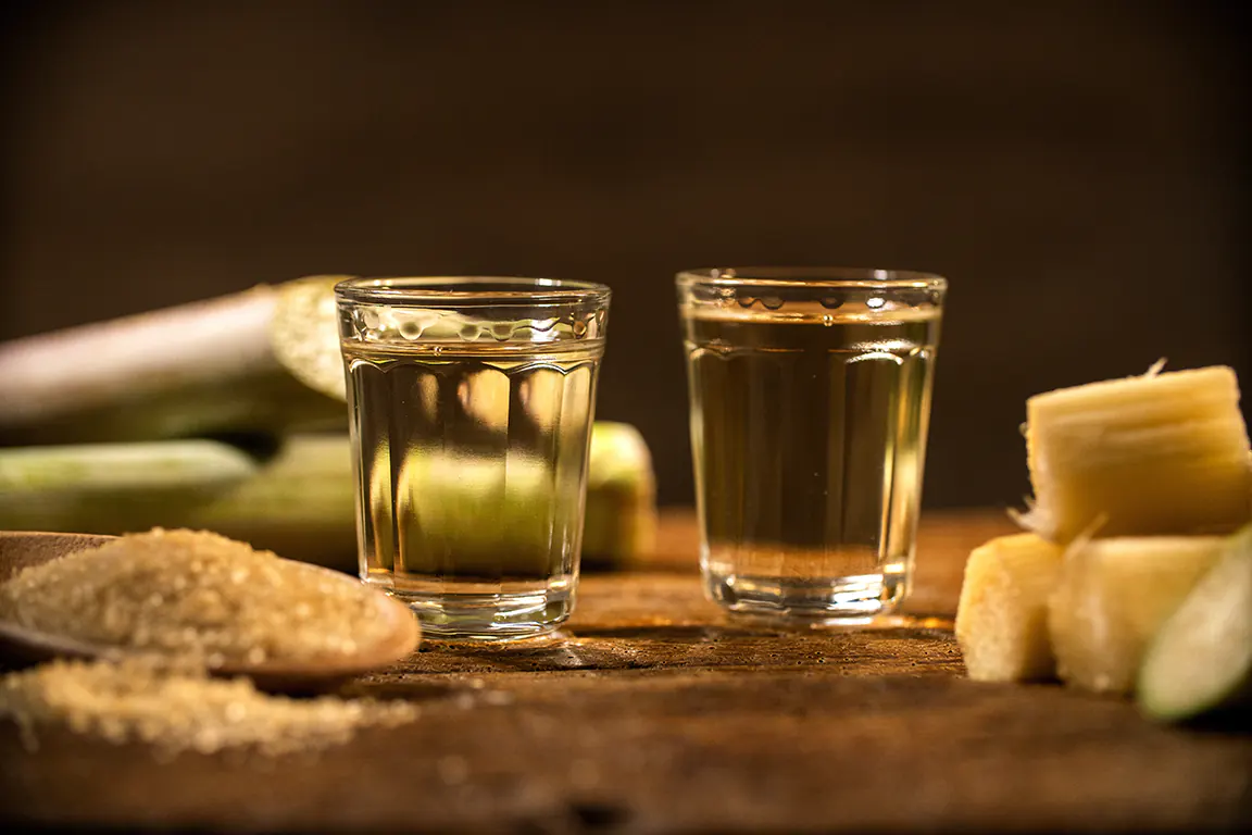 photo d'une table en bois sur laquelle se trouve au premier plan à gauche du sucre de canne en poudre et sur la droite des morceau de cannes à sucrée découpé, au centre de l'image se trouve deux verre rempli de Cachaça, et à l'arrière plan des verres dur la gauche on distingue des tiges de cannes à surexposé sur la table. L'arrière plan est flou, couleur bois et sombre